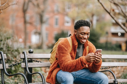 Image of a young man sitting on a bench and smiling at his mobile device