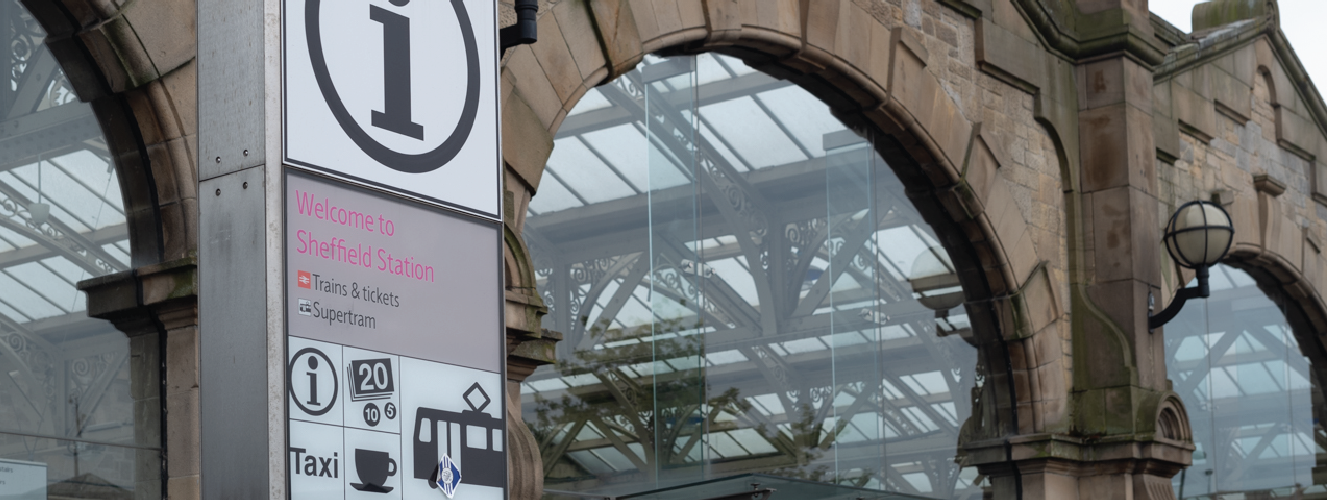 Image shows a tall metal sign displaying a railway symbol, outside of Sheffield train station which is a stone building with large arched glass windows.