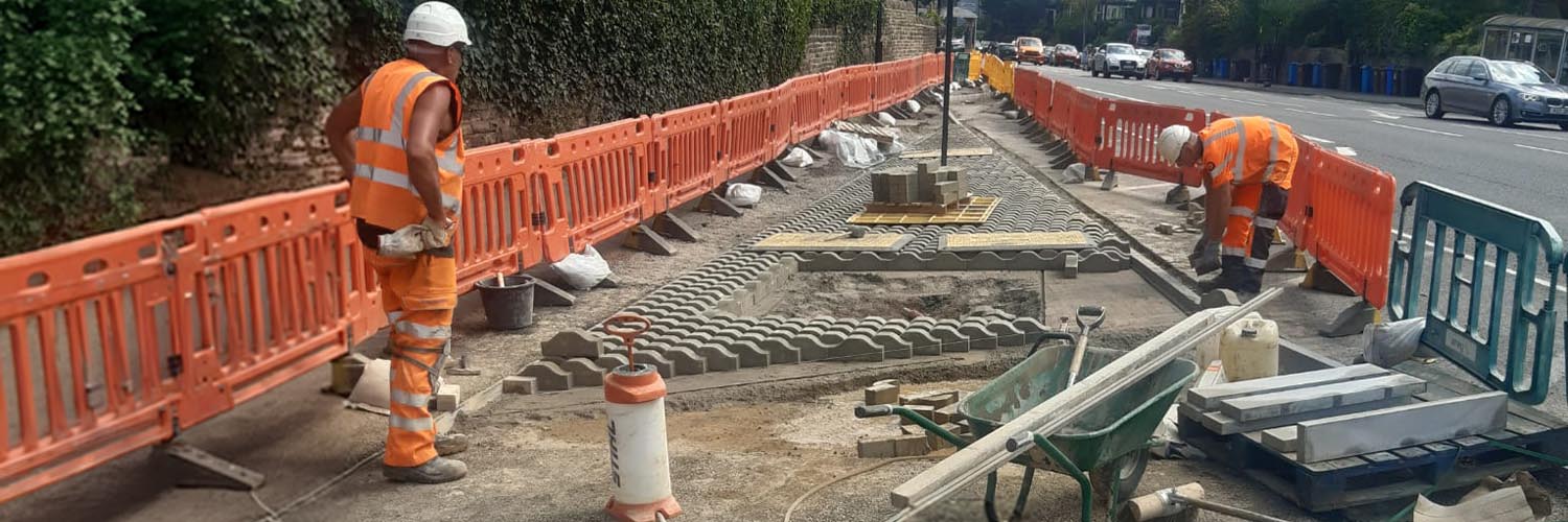 Two construction workers working on roadworks on a pavement beside a residential street.